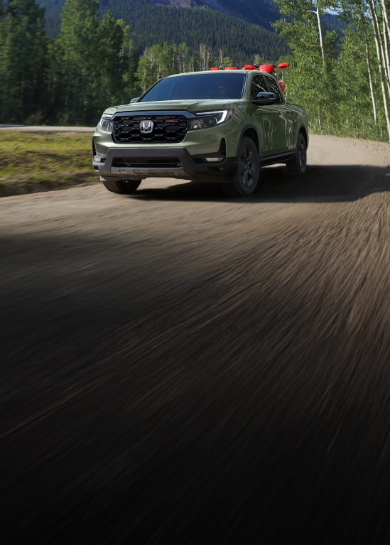 2026 Honda Ridgeline TrailSport in Ash Green Metallic front driving on a scenic dirt road in the mountains (Midsize Pickup Truck).